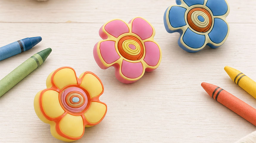 Three flower-shaped children's knobs in pink, yellow, and blue on a light surface with crayons around them.